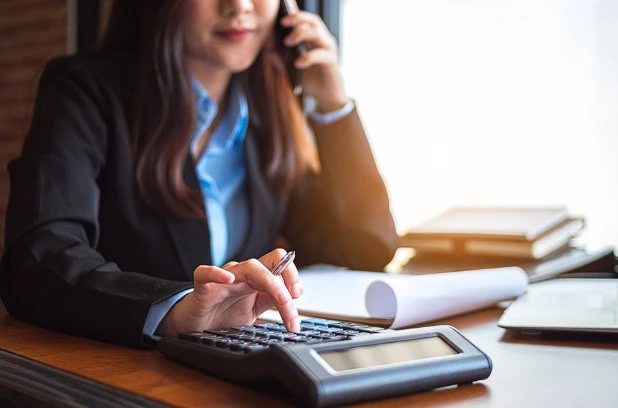 An image of a woman using a calculator while on the phone, estimating the cost to install a furnace and AC.